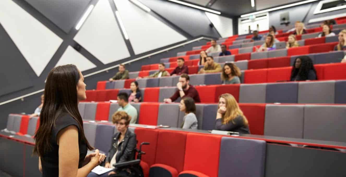 A photo of a lecture hall with a lecturer talking at the front and students sprinkled across the seats which are red and grey