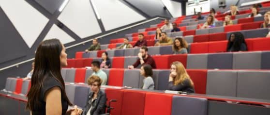 A photo of a lecture hall with a lecturer talking at the front and students sprinkled across the seats which are red and grey