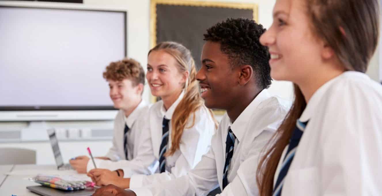 Four students are sat at a table wearing school uniform which is a white shirt with a blue tie. The photo has been taken from a side angle, they are smiling and looking ahead as if they were looking at a teacher off screen.