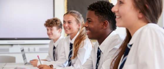 Four students are sat at a table wearing school uniform which is a white shirt with a blue tie. The photo has been taken from a side angle, they are smiling and looking ahead as if they were looking at a teacher off screen.
