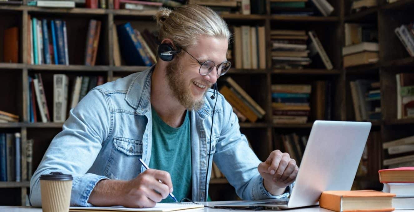 A young man, with blonde hair in a bun, is sat in front of books on shelves as if he were in a library. He is wearing headphones and smiling at his laptop screen as if in conversation