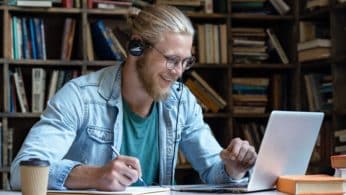 A young man, with blonde hair in a bun, is sat in front of books on shelves as if he were in a library. He is wearing headphones and smiling at his laptop screen as if in conversation