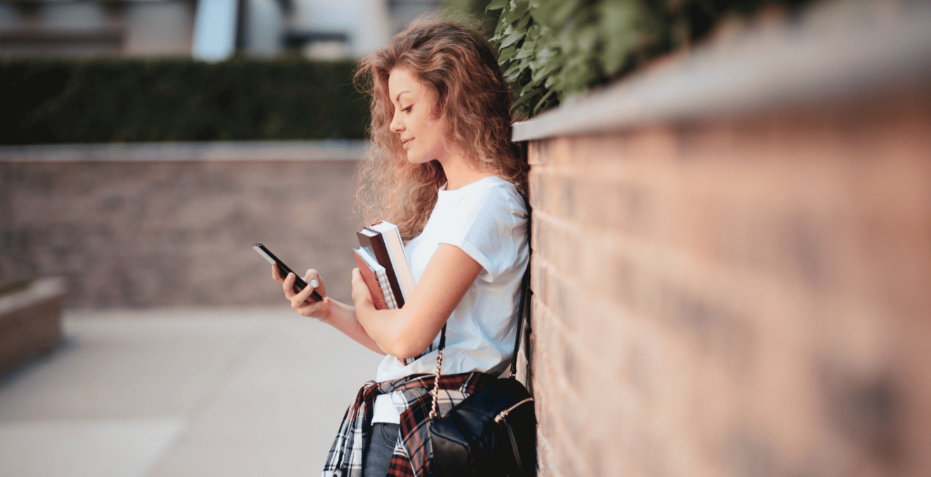 A female student leaning against a wall, looking down at her phone and smiling.