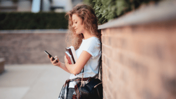 A female student leaning against a wall, looking down at her phone and smiling.