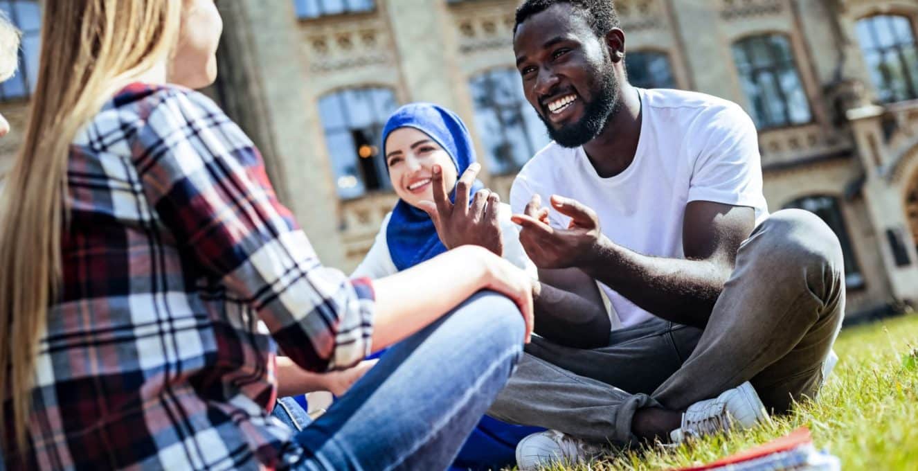 A photo of three students of different ethnicities, sitting on the grass outside of their university building and smiling.
