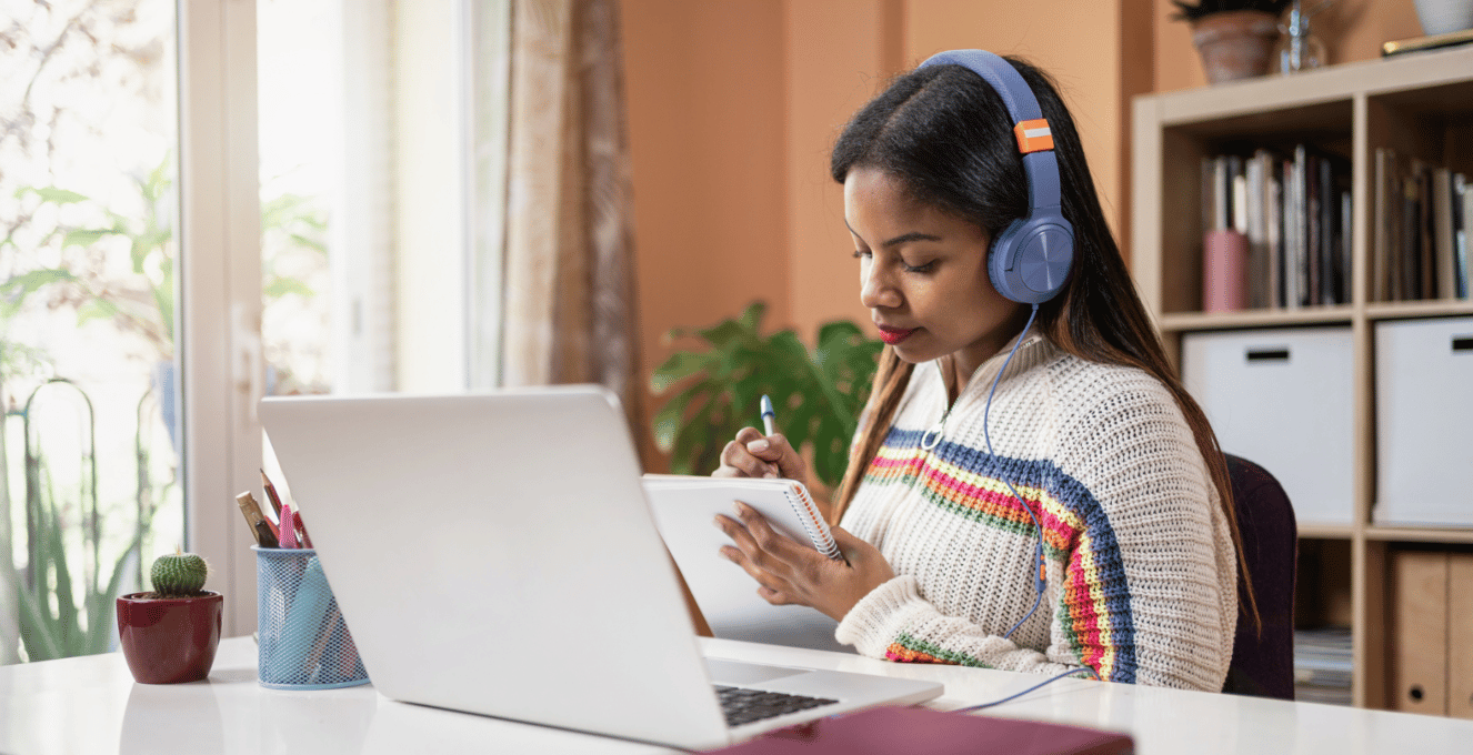 A photo of a young female student wearing headphones and writing on a notepad with her laptop on a desk.