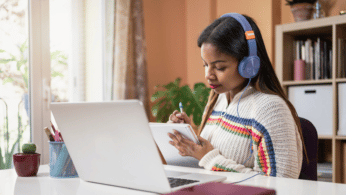 A photo of a young female student wearing headphones and writing on a notepad with her laptop on a desk.