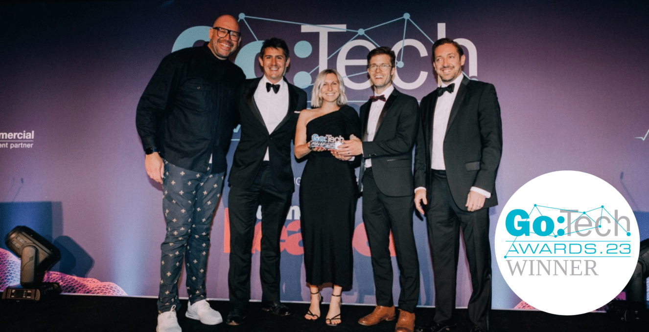A photo of Chris, Claire and Tom at the Go:Tech Awards, standing next to the hosts and smiling for a photo with Claire holding the Go:Tech Awards Trophy. The Go:Tech Awards logo is pictured in the bottom right corner against a white circular background.