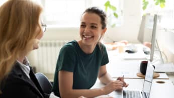 A young female employee smiling at her female boss in an office, with a laptop sitting on the desk in front of them.