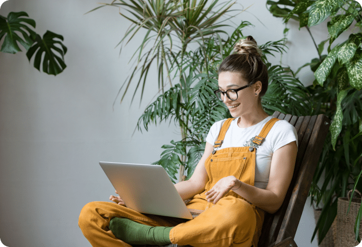 A young female wearing yellow dungarees, sitting on a chair with her laptop next to a plant, smiling.