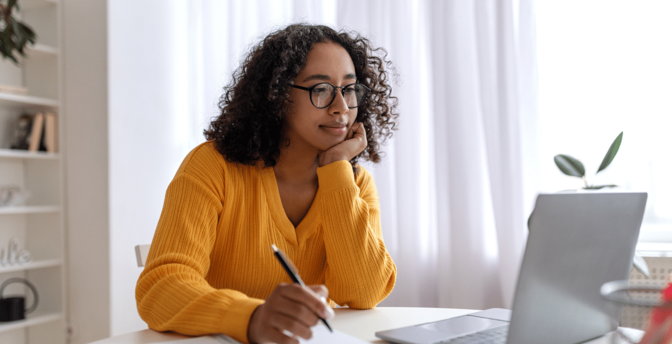 A photo of a young woman sitting at a desk, wearing a yellow jumper and writing down some notes on a notepad.