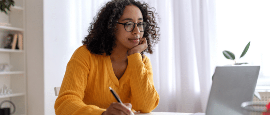 A photo of a young woman sitting at a desk, wearing a yellow jumper and writing down some notes on a notepad.