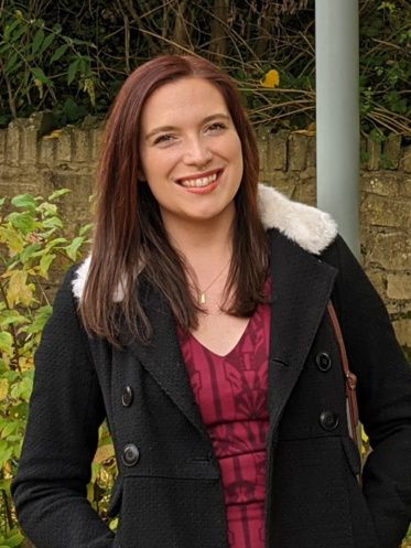 Photo of Gabrielle Wright, she has long brown hair and is wearing a red top and coat with a fluffy hood. She is stood outside with greenery surrounding her and is smiling at the camera.