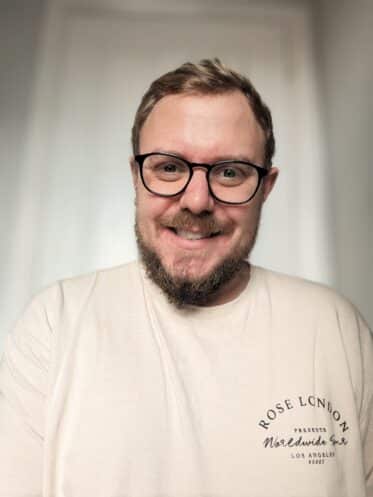 A photo of Daniel Wheeler. He is wearing a cream t shirt, has a beard and dark blonde hair. He is wearing glasses and smiling into the camera.
